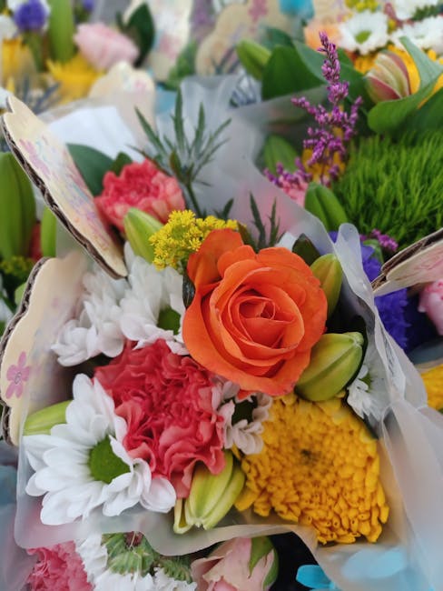A vibrant floral bouquet featuring an orange rose at the center, surrounded by white daisies, pink carnations, yellow chrysanthemums, purple statice, and green foliage. The arrangement is wrapped in c