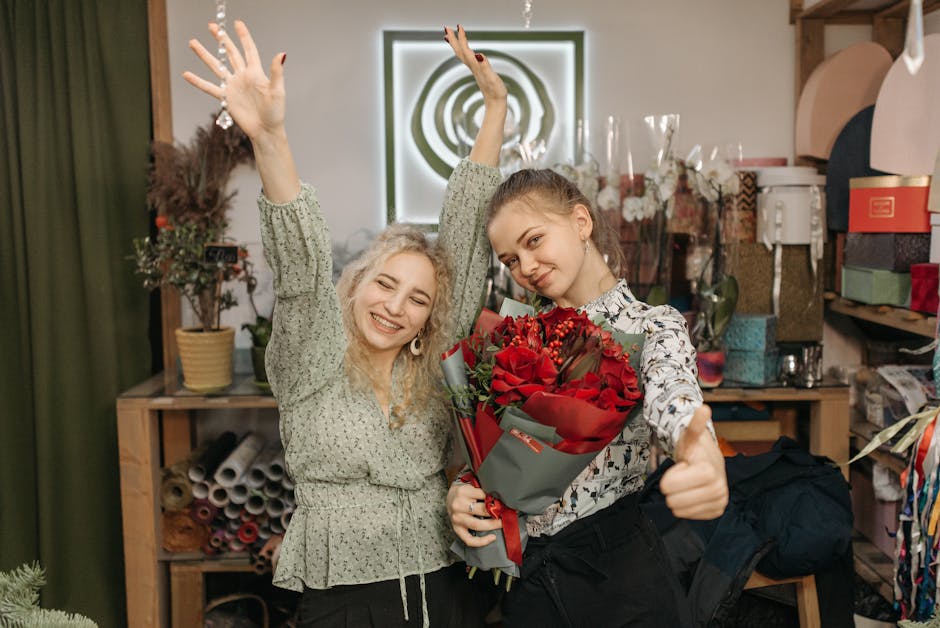 Two young women in a floral shop, with a cozy background of potted plants, wrapping paper rolls, and gift boxes, are celebrating a floral gift delivery. The woman on the right is holding a vibrant bou